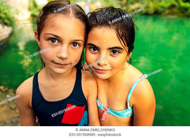 Portrait of a two cute little girls standing near the mountain's lake, with pleasure spending time in summer camp, happy best friends enjoying summer time...