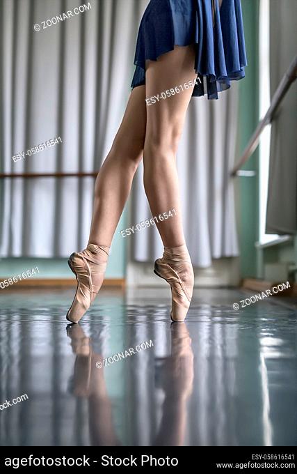 Harmonic legs of a ballet dancer in a blue dance wear who stands on pointes next to the ballet barre. She wears beige pointe shoes