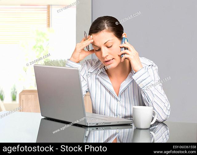 Young woman sitting at desk working with laptop computer at home, talking on mobile phone