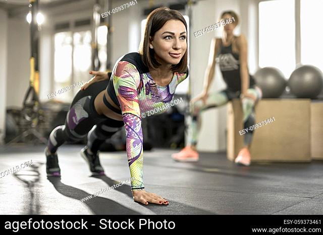 Smiling girl stands in the plank on the right hand in the gym on the windows background. She wears a colorful top with same pants and dark sneakers