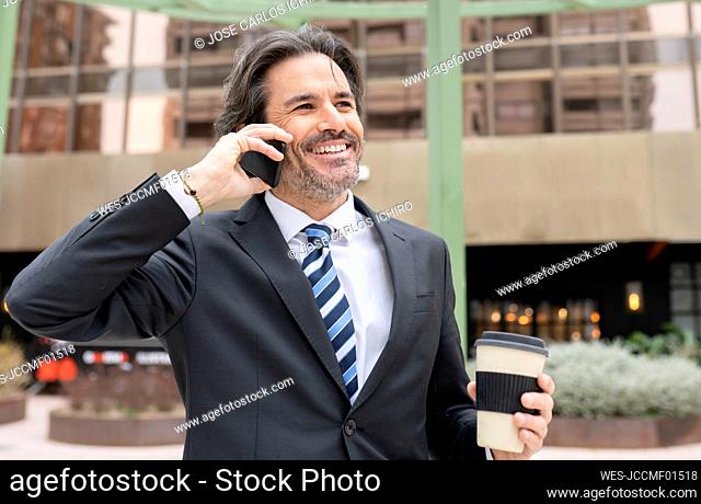 Smiling businessman with coffee cup talking on mobile phone in office park