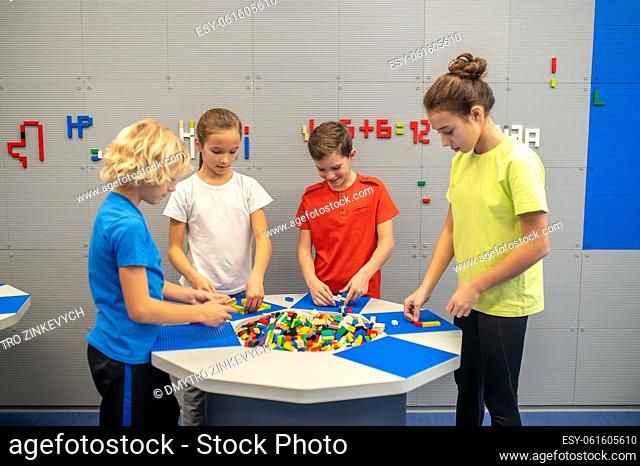 Leisure, game. Happy boys and girls playing with lego standing around table in play area of school