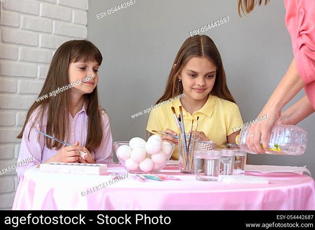 Mom pours vinegar into glasses with food coloring, children at the table prepare to paint eggs