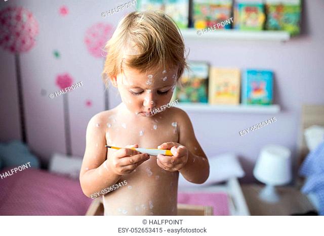 Beautiful little two year old girl at home sick with chickenpox, white antiseptic cream applied to the rash. Holding thermometer, looking at temperature