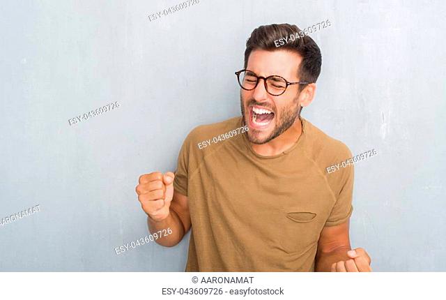 Handsome young man over grey grunge wall wearing glasses very happy and excited doing winner gesture with arms raised, smiling and screaming for success