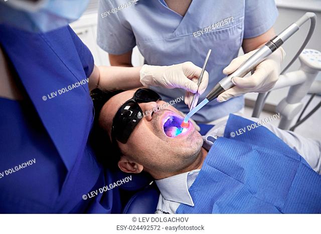 people, medicine, stomatology and health care concept - female dentist and assistant with dental curing light and mirror treating male patient teeth at dental...