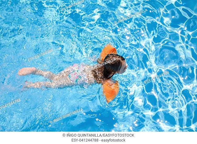 overhead shot of four years old blonde child with orange floater sleeves in arms, armbands, swimming in blue beautiful water of pool