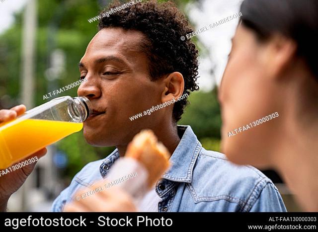 Side view shot of African-American man drinking orange juice and his Latin-American female friend eating a sandwich