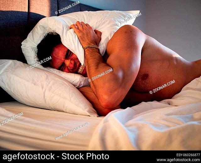 Handsome young male insomniac covering his head and ears, trying to block out the sound with a pillow, as he is kept awake by loud noises around him