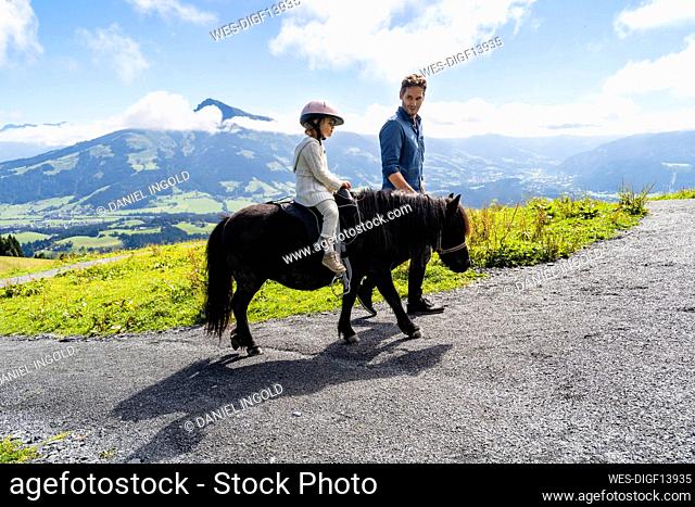 Father walking beside little daughter riding pony against mountain valley