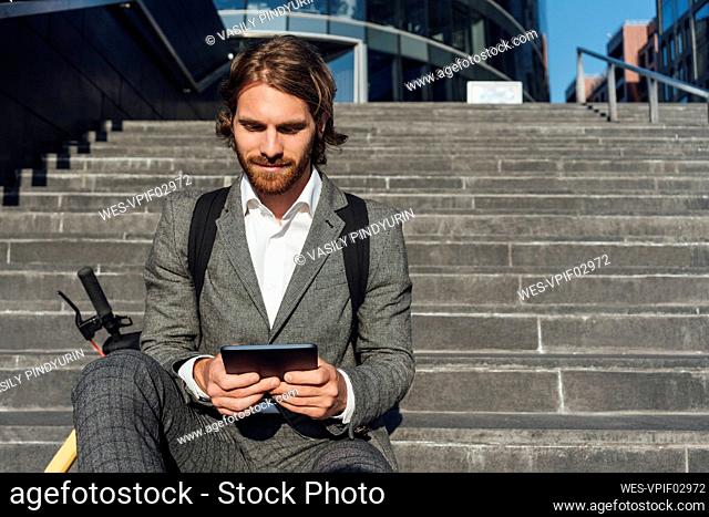 Young handsome male professional using digital tablet while sitting with electric push scooter on steps at downtown