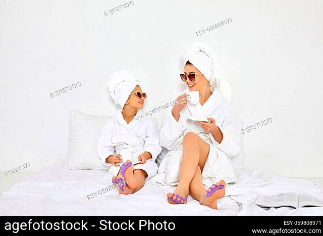 glamorous woman and her daughter in white bathrobes and towels on heads with pedicure sponge on fingers drinking tea