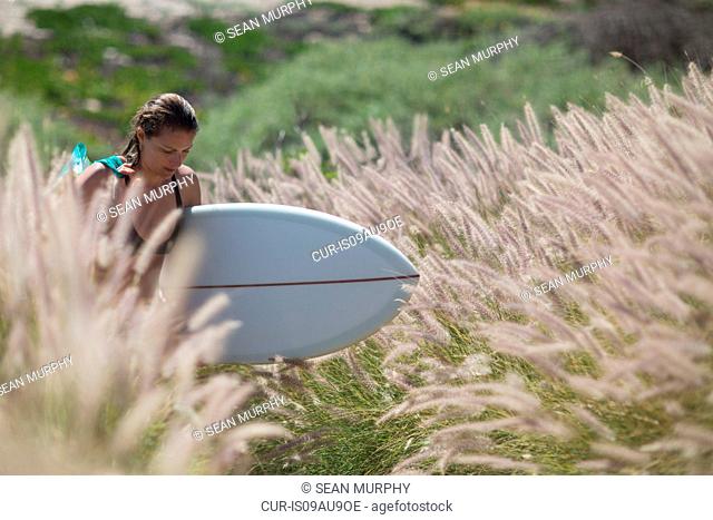 Young woman walking through tall grass carrying surfboard looking down