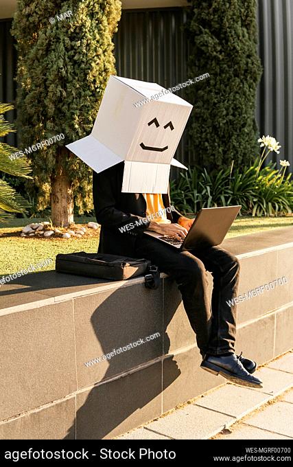 Businessman wearing box with smiley face using laptop sitting on wall