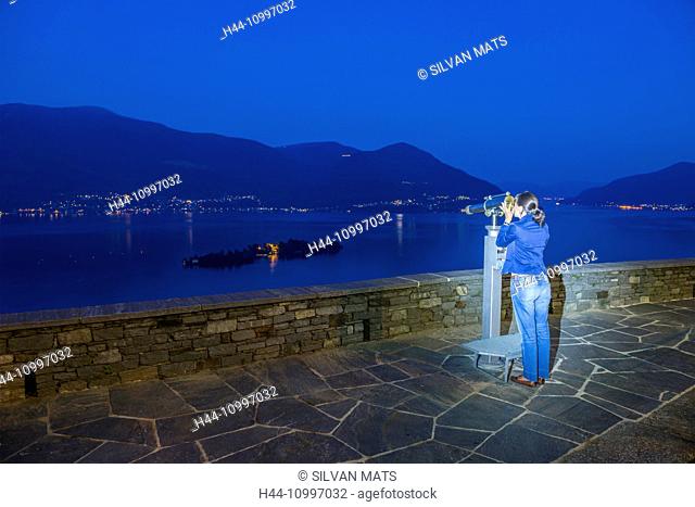 Woman using telescope in blue hour and watching Brissago islands on alpine lake Maggiore with mountain in Ticino, Switzerland