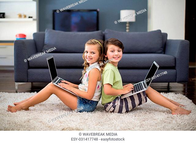 Smiling siblings sitting back to back and using laptop in living room