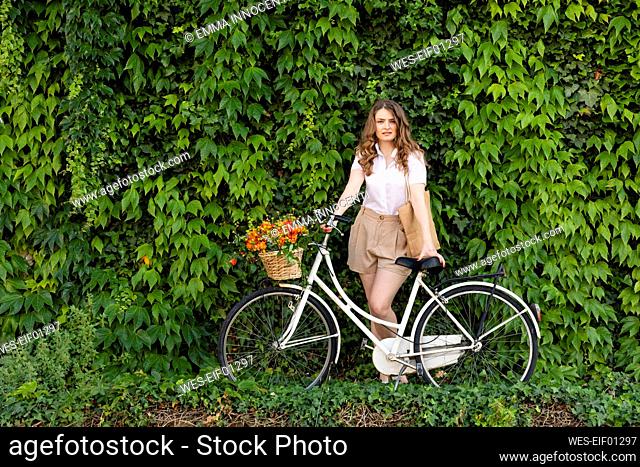 Young woman standing with bicycle in front of green ivy hedge