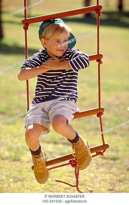 portrait, outdoor, park-scene, blond 7-year-old boy wearing a green baseball-cap sits on a rope-ladder  - GERMANY, 22/08/2004