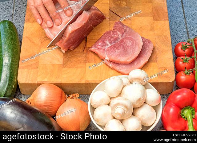 Closeup front view of woman slicing meat on a wooden board. Various types of vegetables and mushrooms are lying on the table