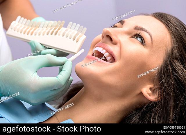 Happy girl in a blue shirt in the dentist's office. Doctor in green gloves makes comparison of the patient teeth with the dental whitening chart