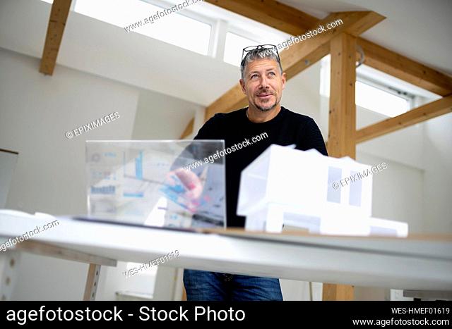 Contemplative architect with futuristic tablet PC and model home standing by desk