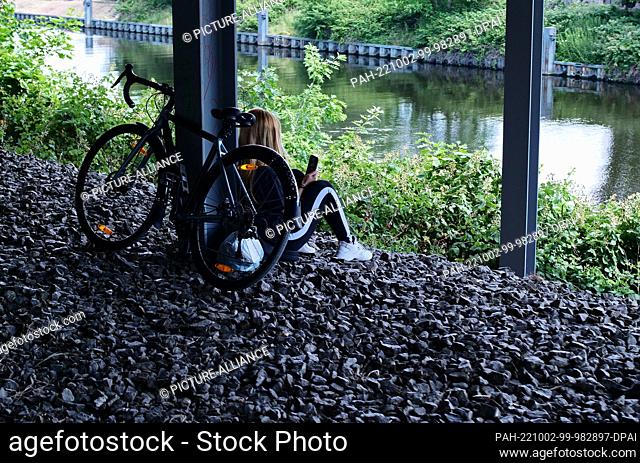26 June 2022, Berlin: 26.06.2022, Berlin. A girl sits alone with her bike under a bridge by a canal, looking at her smartphone