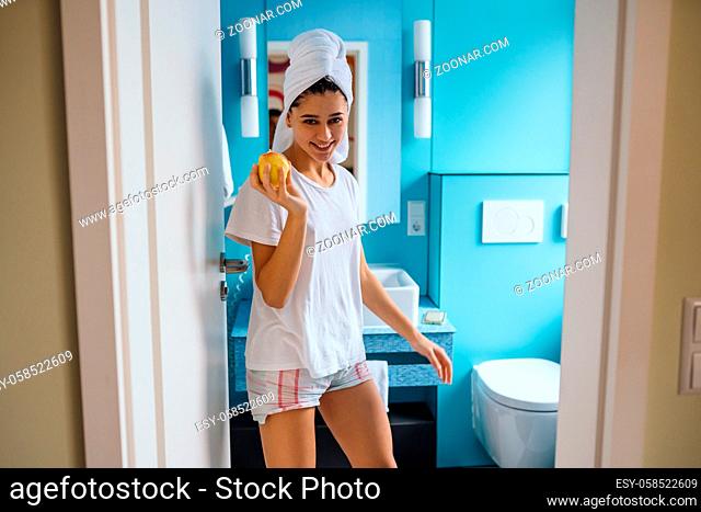 Young caucasian woman wearing towel on head and t-shirt in bathroom, hold apple
