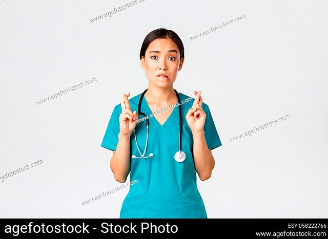 Covid-19, healthcare workers, pandemic concept. Hopeful nervous asian female intern in scrubs waiting for news, doctor cross fingers and biting lip as looking...