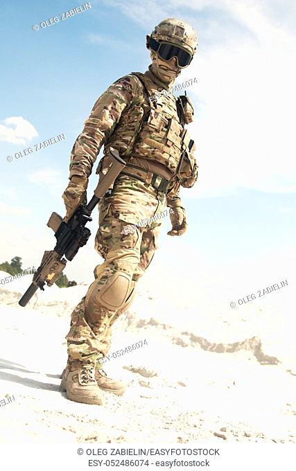 Airsoft player, military games participant in U. S. army infantry camouflage uniform, tactical mask, helmet and glasses, standing in desert area with combat...