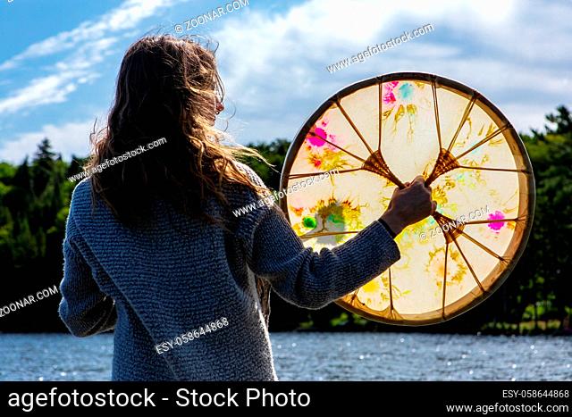 Rear view of young shamanic woman holding sacred native frame drum while in lake praying an worshiping in Northern Quebec in Canada