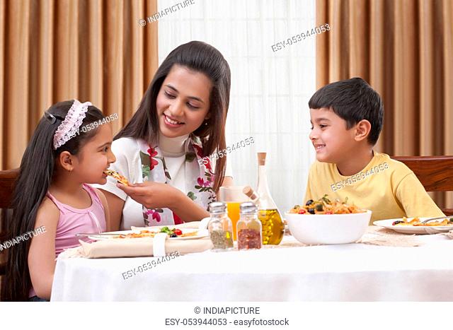 Cheerful mother feeding her daughter pizza with son sitting besides