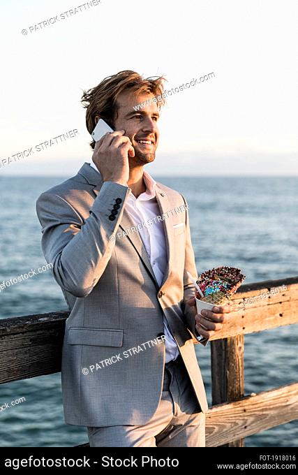 Businessman with ice cream cone talking on smart phone on ocean pier