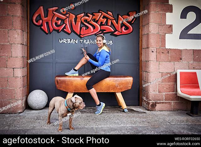 Smiling sportswoman with digital tablet sitting on bench against graffiti door
