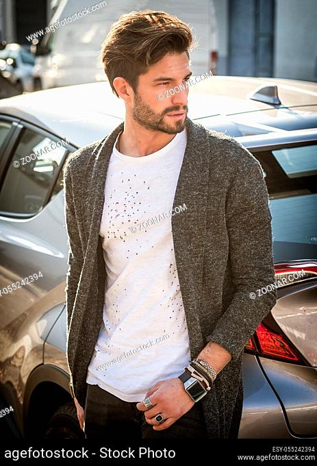 Portrait of young attractive man in white shirt leaning on his new stylish polished car outdoor in city street