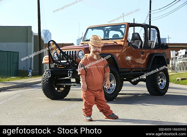 Full length portrait of Caucasian male adult dwarf (little person) standing in street with a off-road vehicle parked behind him
