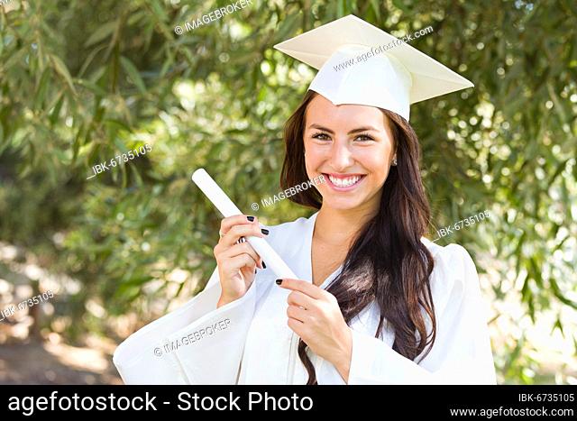 Attractive mixed-race girl celebrating graduation outside in cap and gown with diploma in hand