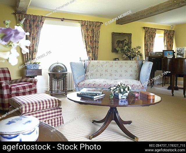 Country cottage sitting room with a beamed ceiling, floral curtains, pedestal coffee table, and an upholstered sofa and chair