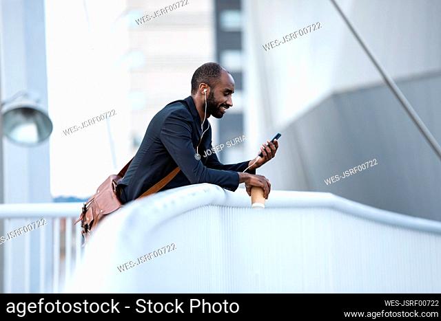Smiling young businessman with earphones and coffee to go looking at smartphone