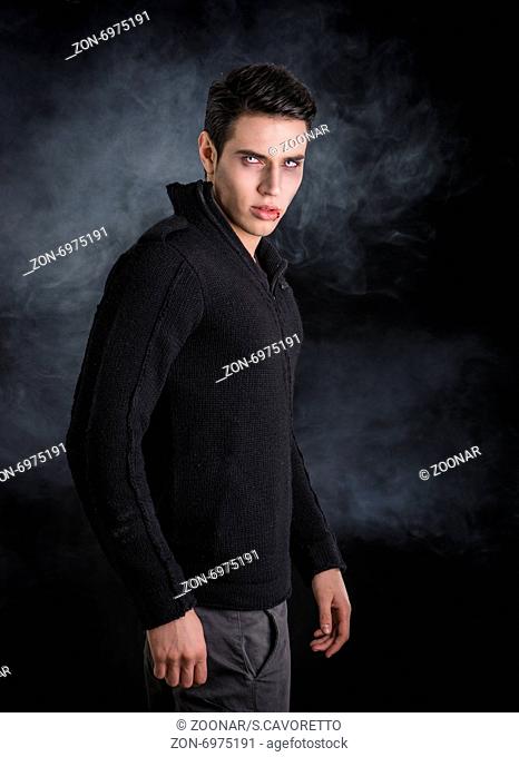 Portrait of a Young Vampire Man with Black Sweater, Looking at the Camera, on a Dark Smoky Background