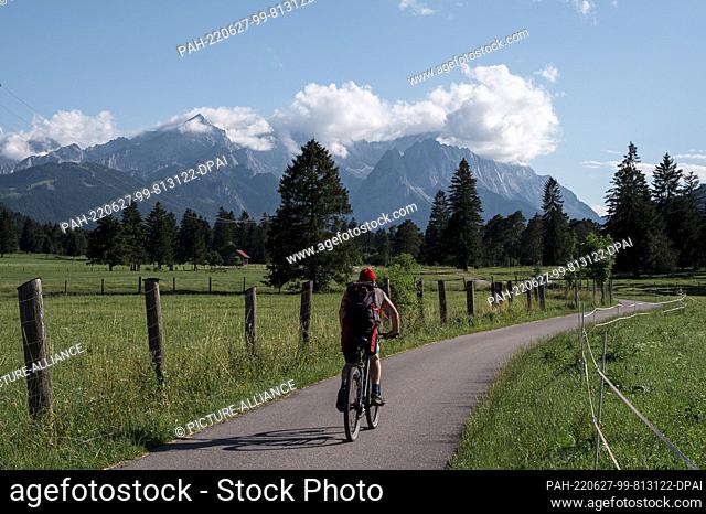25 June 2022, Bavaria, Farchant: A cyclist rides on the bike path between Oberau and Farchant in front of the Wetterstein Mountains