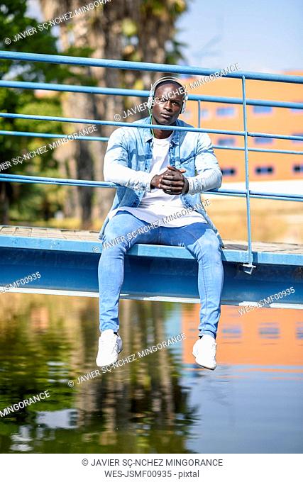 Portrait of man wearing casual denim clothes sitting on footbridge listening music with wireless headphones