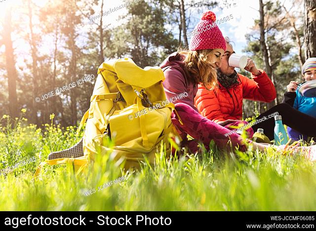 Happy woman with backpack sitting by friends eating and drinking in forest on sunny day