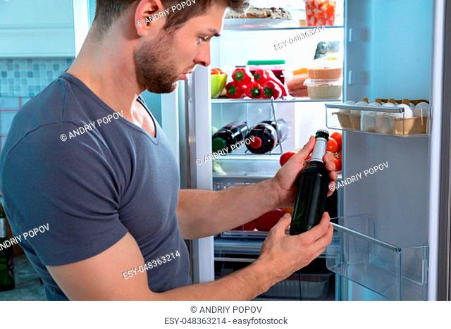 Young Man Holding Beer Bottle Standing In Front Of Refrigerator