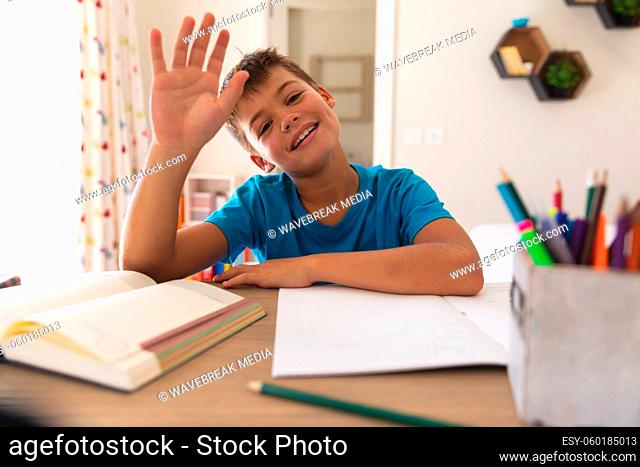 Smiling caucasian boy having video call during class, sitting at desk and waving hand at home
