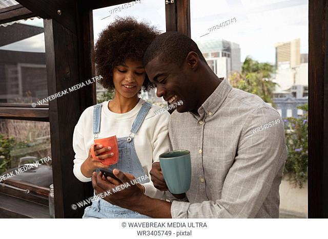 Front view close up of a young mixed race woman and a young African American man standing by a window drinking coffee and looking at his smartphone in a glass...
