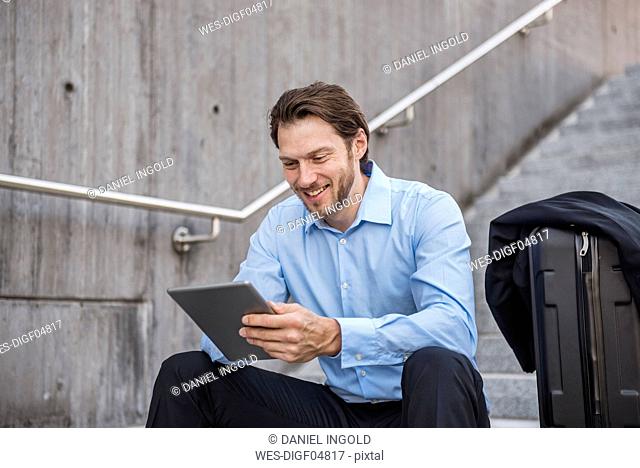 Smiling businessman with rolling suitcase sitting on stairs using tablet