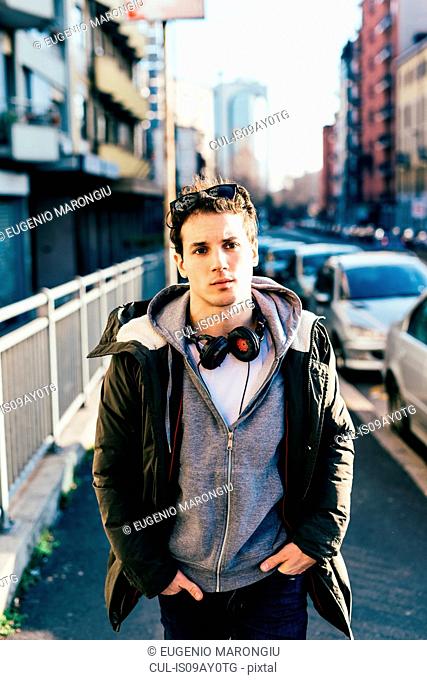 Man with headphones in urban area, hands in pockets looking at camera