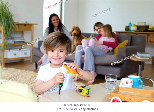 Little boy smiling at the camera while playing with his toy dinosaurs. His family are out of focus in the background