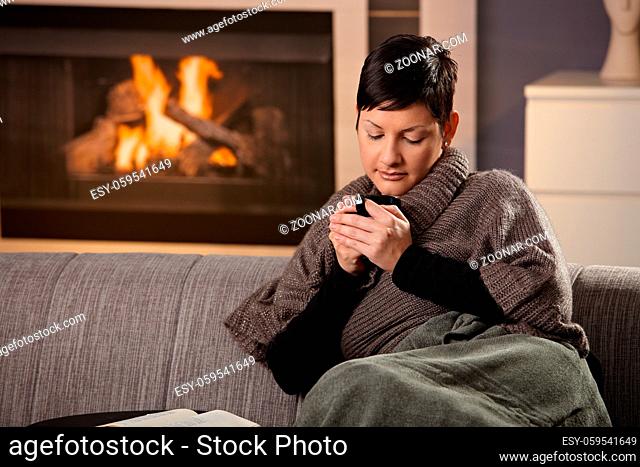 Woman sitting on sofa at home on a cold winter day, drinking hot tea, looking down