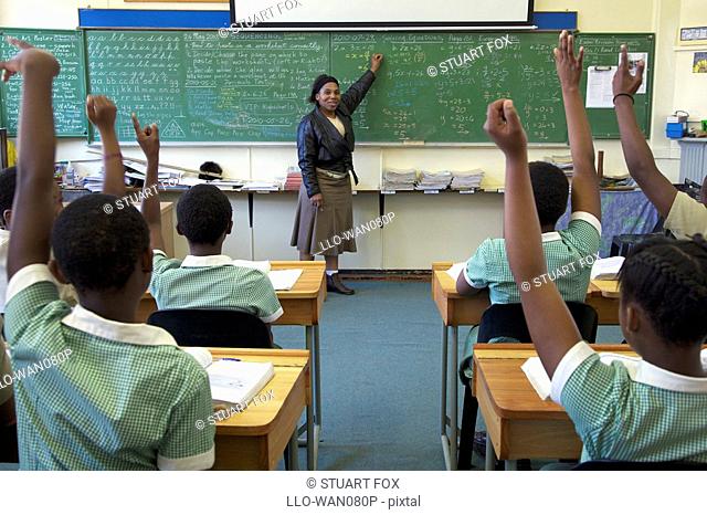 View of a full classroom of pupils with their hands raised, looking toward teacher, KwaZulu Natal Province, South Africa
